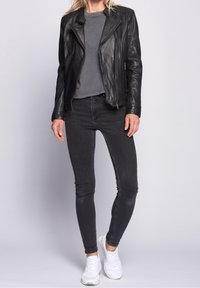 Žena v černé kožené bundě, šedém tričku, tmavých skinny džínách a bílých teniskách stojí proti jednoduchému světlému pozadí.