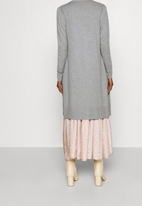 Langer grauer Cardigan aus Strickmaterial, mit geradem Schnitt und langen Ärmeln, getragen über einem rosa Blumenrock und beigen Ankle Boots.