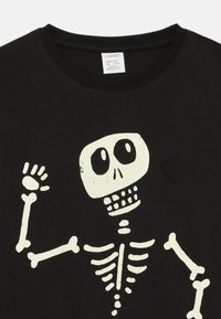 Black cotton t-shirt featuring a white skeleton graphic, with a large skull, hand raised, and clear bone details. Tag visible on the collar.