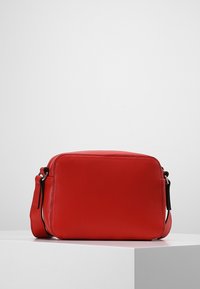 Červená kožená taška přes rameno s obdélníkovým tvarem, hladkým povrchem, zipovým uzávěrem a nastavitelným popruhem. Doplňky zahrnují černé kovové prvky.