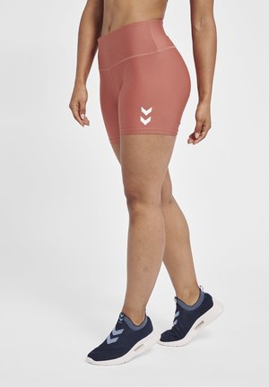 Henkilö, jolla on päällään korkeavyötäröiset tomunpunaiset pyöräilyshortsit, joissa on valkoiset shevron-logot, sekä tummansiniset slip-on-urheilukengät, joissa on valkoiset pohjat.