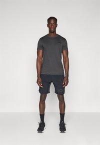 Tricou sportiv gri asortat cu pantaloni scurți negri cu un logo, completat cu șosete negre și pantofi de alergare. Material neted, design ajustat.