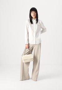 Witte blouse met lange mouwen en een kraag, gecombineerd met lichtbeige wijde broek. Houdt een kleine crèmekleurige tas met een kettinghengsel vast.