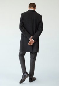 Donkergrijze wollen overjas met een rechte snit, gecombineerd met slanke grijze broeken. Gladde leren schoenen maken de outfit compleet, gezien vanuit de achterkant.