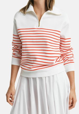 Woman wearing a white long-sleeve pullover with red horizontal stripes and a half-zip collar, paired with a white pleated skirt.