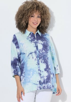 Frau mit lockigem Haar, die ein blau-weißes Batik-Knopfhemd und weiße Hose trägt, vor einem schlichten weißen Hintergrund steht.