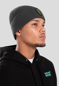 Mladý muž nosící šedou žebrovanou čepici s malou zelenou nášivkou mimozemšťana a černou mikinu s nápisem "Wrong Friends" v tyrkysové barvě.