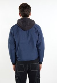 Giacca bomber color navy con cappuccio grigio, design casual, polsini e vita elasticizzati. Presenta una tasca laterale con patta sul braccio sinistro.