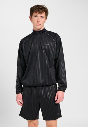 Młody mężczyzna w czarnej bluzie z długim rękawem i suwakiem do ćwiartki oraz dopasowanych szortach z subtelnymi pionowymi paskami i wzorami w formie jodełki na rękawach.