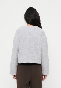 Femme aux longs cheveux foncés portant un pull gris clair en matière pelucheuse à manches longues et un pantalon brun foncé, dos à la caméra.