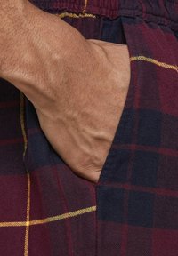 Pantalones cortos de cuadros con una combinación de colores burdeos y azul marino con acentos amarillos. Primer plano de una mano en el bolsillo lateral, mostrando la textura de la tela.