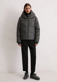 Grijze pufferjack met een brede capuchon, ritssluiting en frontzakken. Heeft ribgebreide manchetten en een boxy silhouet. Draagt bij zwart broek en schoenen.