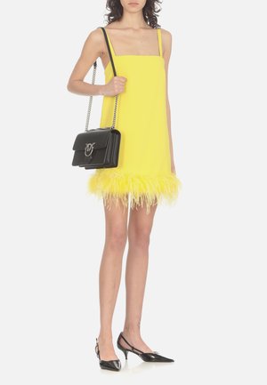 Robe sans manches jaune avec un décolleté carré et un ourlet orné de plumes. Associée à un sac à main noir avec une bandoulière en chaîne et des détails en argent.