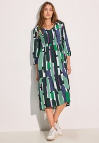 Mekko, jossa on geometrinen kuvio vihreänä, purppurana ja mustana; polvipituinen ja kolmanneksi vartta peittävin hihoin; valmistettu kevyestä kankaasta.