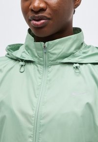 Κοντινό πλάνο ατόμου που φοράει ανοιχτό πράσινο μπουφάν Nike με φερμουάρ, κουκούλα και ρυθμιζόμενα κορδόνια.