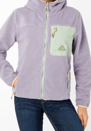 Henkilö, joka on pukeutunut vaaleanpunaiseen fleecetakkiin, jossa on vaaleanvihreä vetoketjullinen rintatasku ja siniset farkut, käsi sivutaskussa.