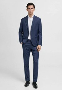 Marineblauer Anzug mit maßgeschneiderter Passform, bestehend aus einem Ein-Knopf-Jackett und flachen Hosen. Kombiniert mit einem weißen Hemd und schwarzen Schuhen.