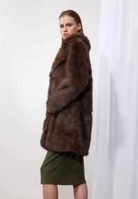 Manteau en fausse fourrure marron avec un col haut, des manches longues et une coupe ample. Porté sur une jupe verte olive arrive au genou. Texture lisse.