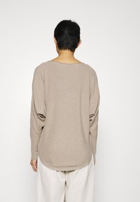 Beige oversized topp med vid halsringning och sänkta axlar, med en texturerad finish och rundad nederkant. Matchad med ljusa byxor.