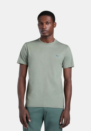 Uomo che indossa una maglietta verde chiaro a maniche corte con un piccolo logo di cane blu sul petto e pantaloni verdi coordinati, in piedi contro uno sfondo bianco.