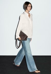 White long-sleeve button-up shirt, light blue flared jeans, and a woven dark brown handbag with a strap. Model wearing flip-flops.