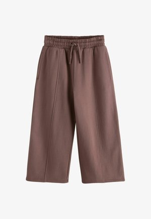 Ung pige med langt mørkt hår iført en hvid langærmet cropped top, løse lilla bukser og hvide sneakers, stående mod en ensfarvet baggrund.