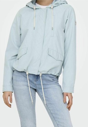 Osoba ubrana w jasnoniebieską kurtkę z kapturem, kieszeniami z przodu i ściągaczami, w połączeniu z jasnoniebieskimi dżinsami na jednolitym tle.