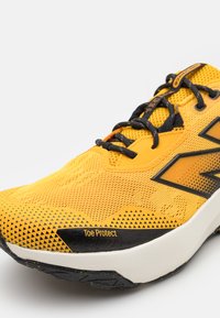 Gul atletiksko med mesh-overdel, sorte akcenter, en tekstureret sål og "Toe Protect" label på siden. Letvægtsdesign.