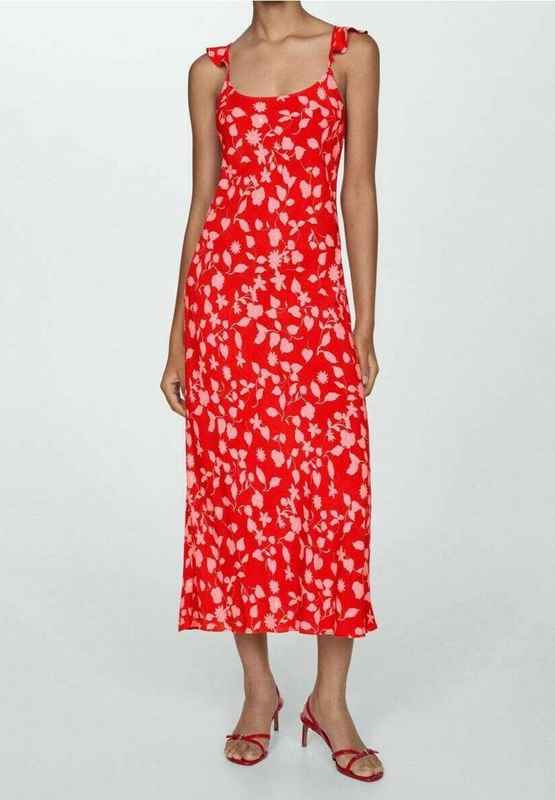 Femme portant une robe midi rouge sans manches avec un imprimé floral rose clair et des sandales à talons rouges assorties, debout devant un fond uni.