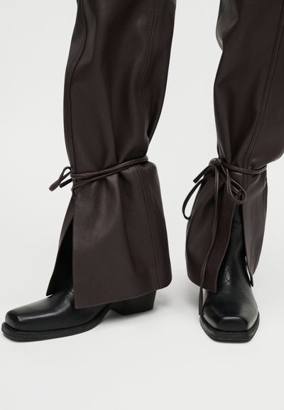 Σκούρο καφέ δερμάτινο παντελόνι με κορδόνια στους αστραγάλους, φορεμένο πάνω από μαύρες δερμάτινες μπότες με τετράγωνη μύτη και χαμηλό τακούνι, ενάντια σε ανοιχτό φόντο.