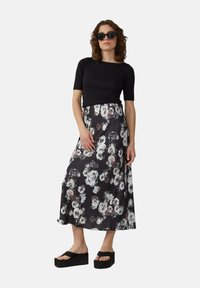 Haut noir à manches courtes associé à une longue jupe florale avec des roses blanches et grises sur un fond noir. Le mannequin porte des sandales à plateforme noires.