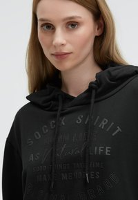 Black hooded sweatshirt with textured print featuring brand name and phrases. Drawstrings are black; fabric appears soft and casual.