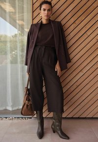 Brauner maßgeschneiderter Blazer über einem dunklen Oberteil, hochtaillierte dunkle Hose, kniehohe olivgrüne Stiefel und eine braune Schultertasche mit Schnalle.