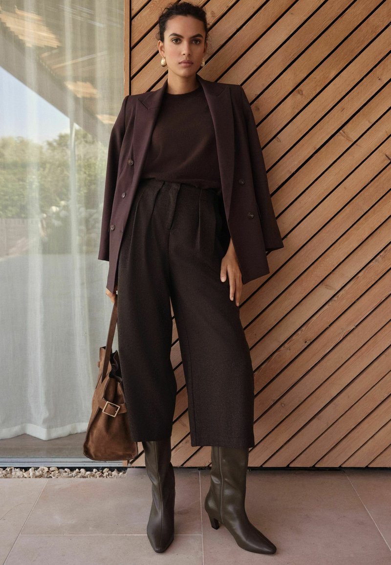 Brauner maßgeschneiderter Blazer über einem dunklen Oberteil, hochtaillierte dunkle Hose, kniehohe olivgrüne Stiefel und eine braune Schultertasche mit Schnalle.