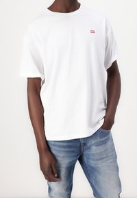 Person som bär en enkel vit Diesel T-shirt och blå jeans, med ena handen i fickan, mot en enkel vit bakgrund.