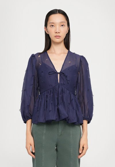 Dunkelblaue Bluse aus transparenter Stoff, mit V-Ausschnitt, Puffärmeln und gerafftem Peplum. Blumenstickereien im gesamten Design. Verstellbarer Bindegürtel vorne.