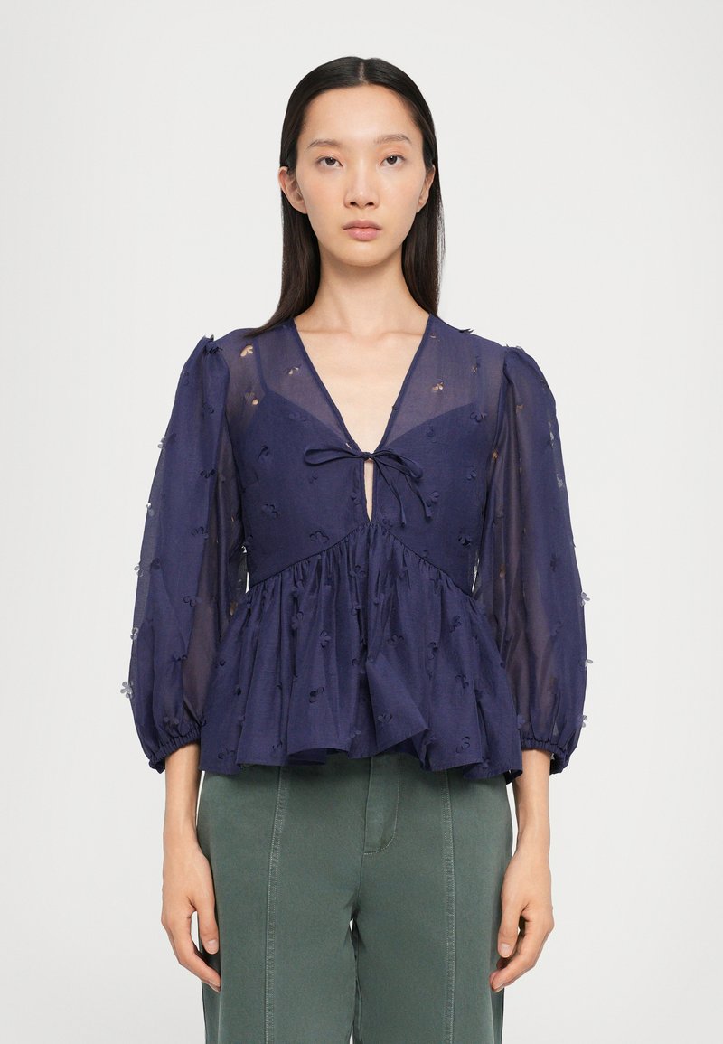 Blusa de color azul marino con tejido transparente, escote en forma de V, mangas abullonadas y peplum fruncido. Detalles de bordado floral por toda la prenda. Cinta ajustable en la parte frontal.