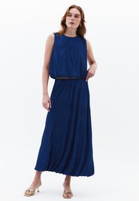 Mouwloze blauwe jurk met een textuur, geplooid ontwerp, met een losse top en een elastische taille. Gecombineerd met gouden sandalen met bandjes.
