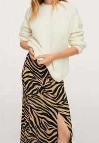 Cremfarbener gestrickter Pullover mit aufgerollten Ärmeln, kombiniert mit einem schwarz-beigen, zebra-streifenförmigen Rock mit einem hohen Schlitz bis zum Oberschenkel.