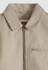 Μπεζ τζάκετ Levi's με μυτερό γιακά, κλείσιμο με φερμουάρ μπροστά και τσέπη στο στήθος με φερμουάρ στη δεξιά πλευρά.