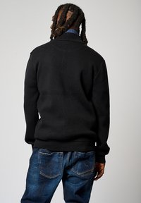 Pull en laine noir avec col châle, associé à un jean bleu foncé, présentant un tissu texturé et des poignets côtelés à la taille.