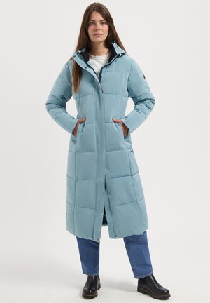 Vrouw staat met een lange lichtblauwe gewatteerde jas aan, met handen in de zakken, blauwe jeans en zwarte enkellaarsjes op een witte achtergrond.