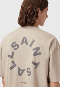 Beige pamut póló, nagy kör alakú grafikával, amelyen a "SAINT" és a "LONDON" sötétszürke felirattal szerepel, rövid ujjakkal és laza szabással.