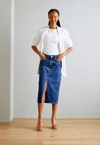 Weißer Blazer über einem figurbetonten weißen Top, kombiniert mit einem knielangen, dunklen Jeansrock mit einem Schlitz vorne und sandfarbenen Plateausandalen.