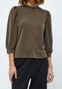 Olivgrön blus med hög halsringning, puffärmar i tre kvartar och en slät textur, kombinerad med svartabyxor.