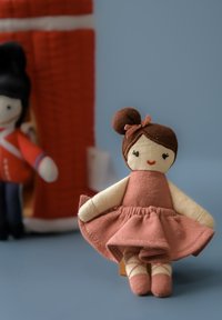 Miękka lalka z brązowym kokiem, ubrana w różową sukienkę i kremową podkoszulkę. Charakterystyczne rumieńce na policzkach oraz czerwone buty z krzyżującym się wzorem.