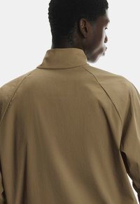 Braune, leichte Jacke mit Stehkragen, raglanÄrmeln und strukturiertem Stoff, von hinten gezeigt.