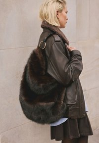 Jachetă maro din piele cu guler din blană de miel purtată peste o cămașă albastră. O geantă mare, pufoasă, din blană maro închis, cu un inel auriu decorativ, este atârnată peste umăr.