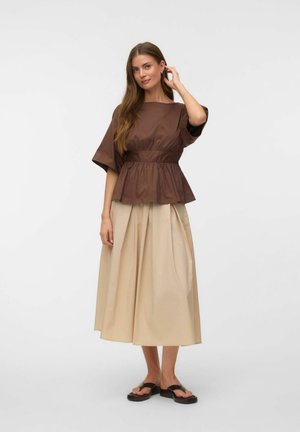 Femme aux longs cheveux bruns portant un haut marron ample à manches courtes et une large jupe midi beige, debout avec une main près de l'oreille.
