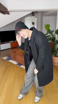 Casaco longo preto, calças cinzentas de perna larga e ténis prateados. Visual em camadas com gorro preto. Ambiente interior iluminado com pavimento em madeira.